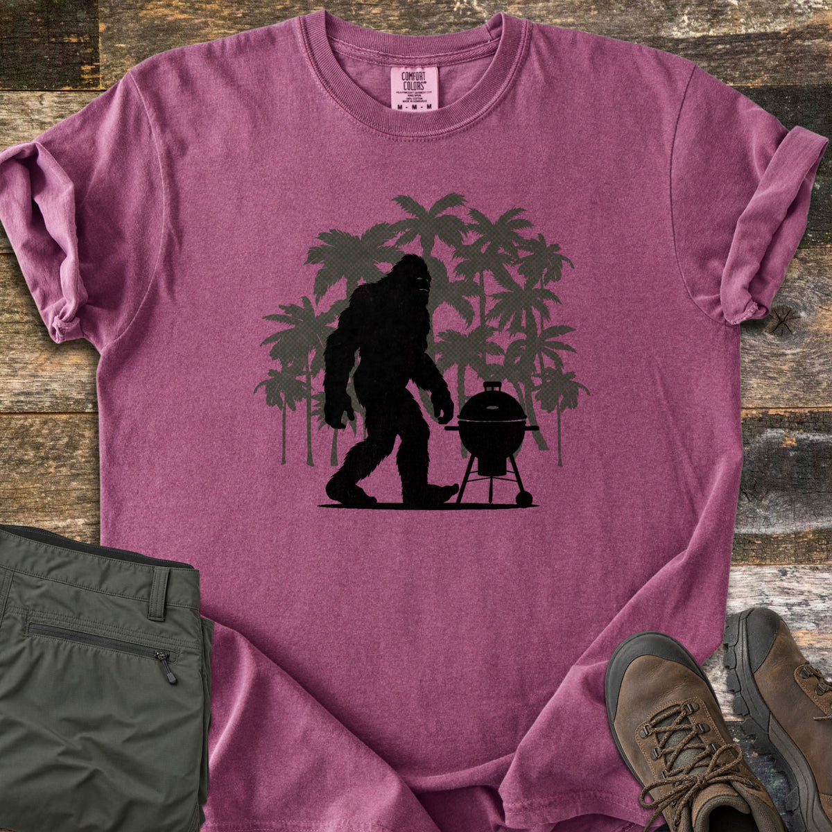 Squatch BBQ Smoker Comfort Colors T-shirt in Berry, laid flat. A black Bigfoot silhouette walks past a kettle grill on wheels, set against dark green palm tree silhouettes; no words are printed. The shirt lies on weathered wood beside olive pants and brown hiking boots.