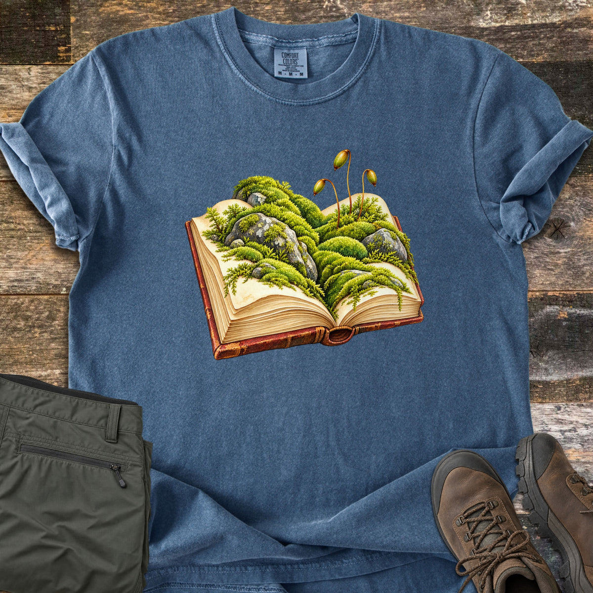 Moss Storybook Comfort Colors T-shirt in Denim, laid flat. An open antique book with a red cover has green moss, gray rocks, and moss spore stalks growing from its pages. No words are printed. It lies on weathered wood beside olive hiking pants and brown hiking boots.
