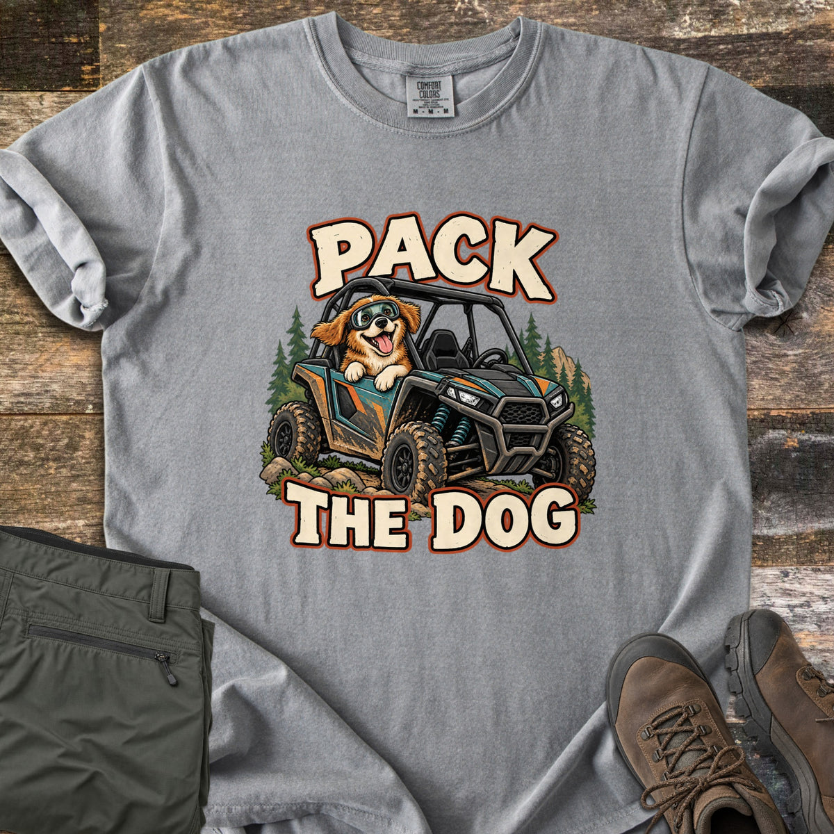 Pack the Dog Comfort Colors T-shirt in Grey, laid flat. A cartoon dog in goggles rides in a muddy off-road side-by-side among pine trees, between the words Pack The Dog. Sleeves rolled, on weathered wood beside olive hiking pants and brown hiking boots.