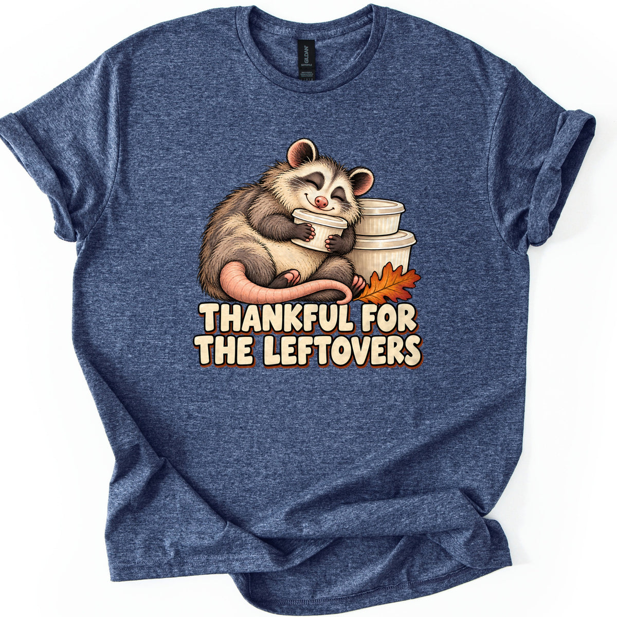 Thankful For The Leftovers Comfort Colors T-shirt in Heather Navy, laid flat. A chubby opossum smiles with eyes closed while hugging a white food container by a stack of containers and an orange leaf, over the words Thankful For The Leftovers. Sleeves are rolled on a white surface.
