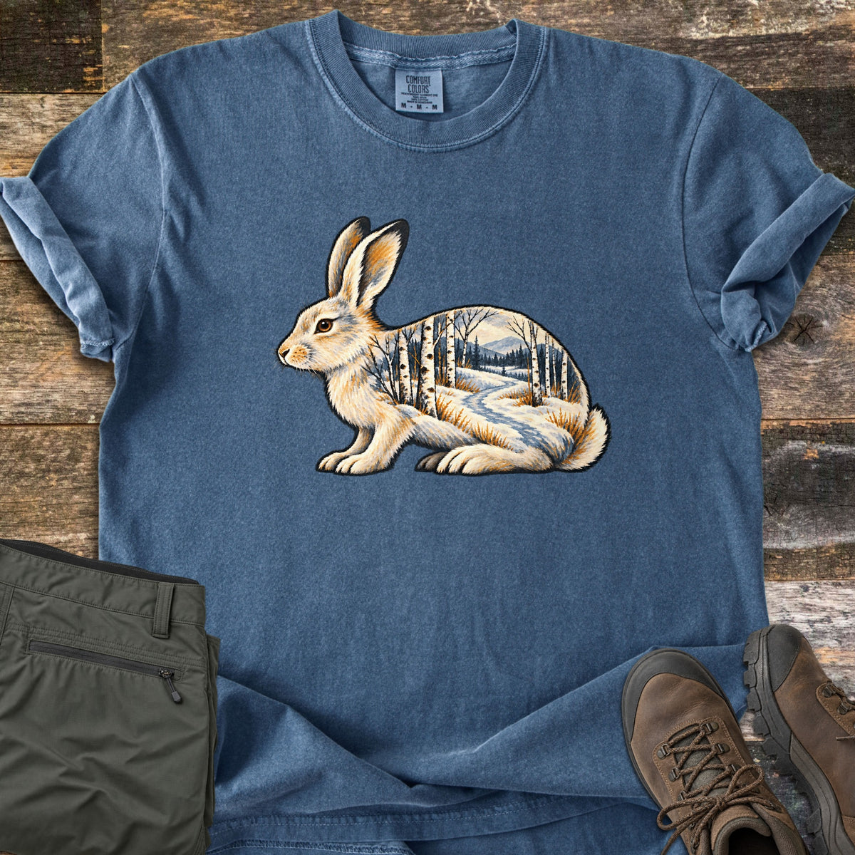 Snowshoe Hare Birch Woods Comfort Colors T-shirt in Denim, laid flat. A white snowshoe hare sits in profile, its body filled with a snowy birch forest, a winding path and distant mountains. Olive hiking pants and brown boots lie beside it on weathered wood planks.
