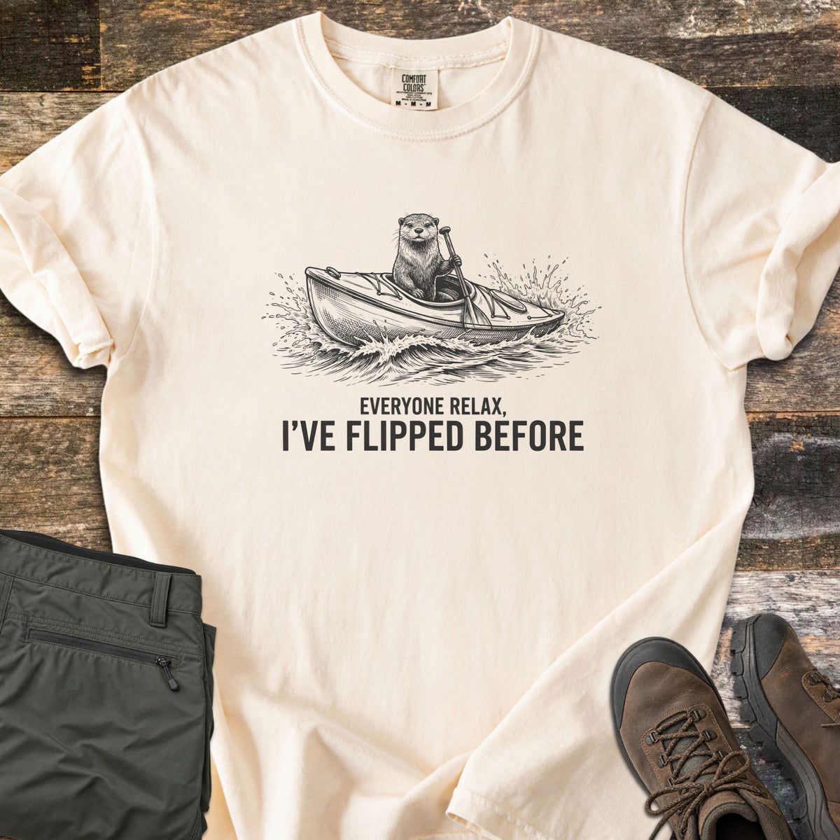 Everyone Relax I've Flipped Before Comfort Colors T-shirt in Ivory, laid flat. A black ink sketch of an otter paddling a kayak through splashing waves above the words Everyone Relax, I've Flipped Before. Folded olive hiking pants and brown boots lie beside it on weathered wood.