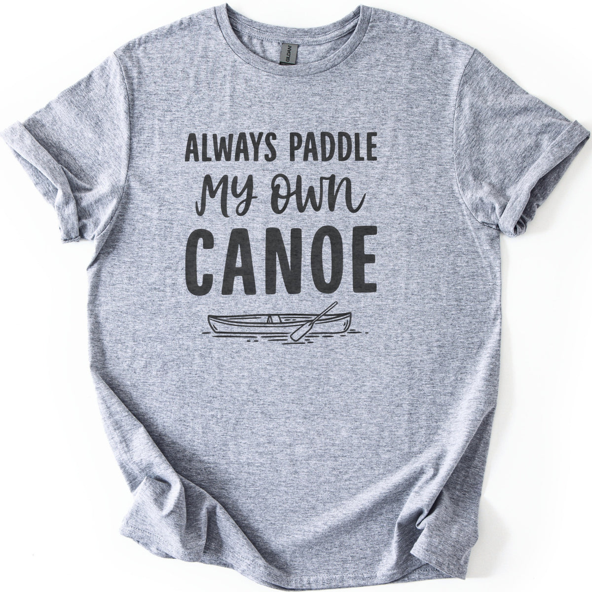Paddle My Own Canoe Comfort Colors T-shirt in Sport Grey, laid flat. Dark charcoal text reads Always Paddle My Own Canoe above a line drawing of a canoe with a paddle on rippling water. The shirt has rolled sleeves and lies on a white surface.