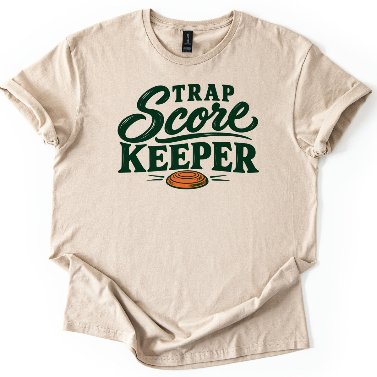 Trap Score Keeper Comfort Colors T-shirt in Sand, laid flat. Dark green lettering reads Trap Score Keeper, with Score in flowing script above an orange clay target with motion lines. The sleeves are cuffed and the hem is loosely folded on a plain white background.
