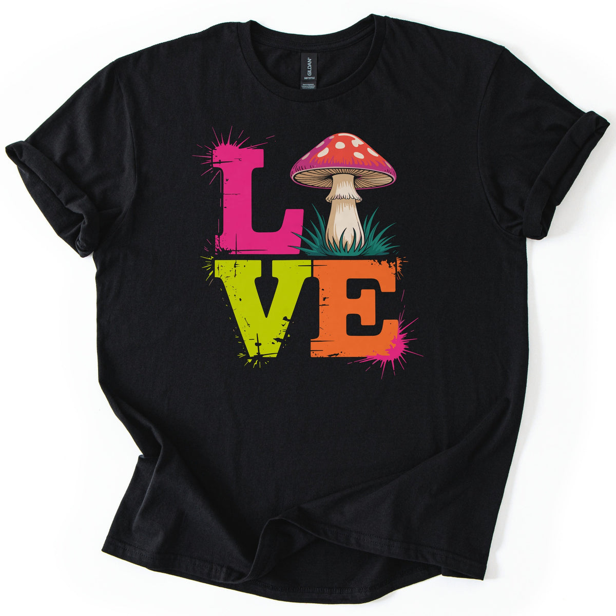 Mushroom Love Comfort Colors T-shirt in Black, laid flat. Stacked distressed letters spell LOVE: a pink L, a red mushroom with white spots standing in green grass as the O, a lime green V and an orange E, with paint splatters. Cuffed sleeves on a white background.