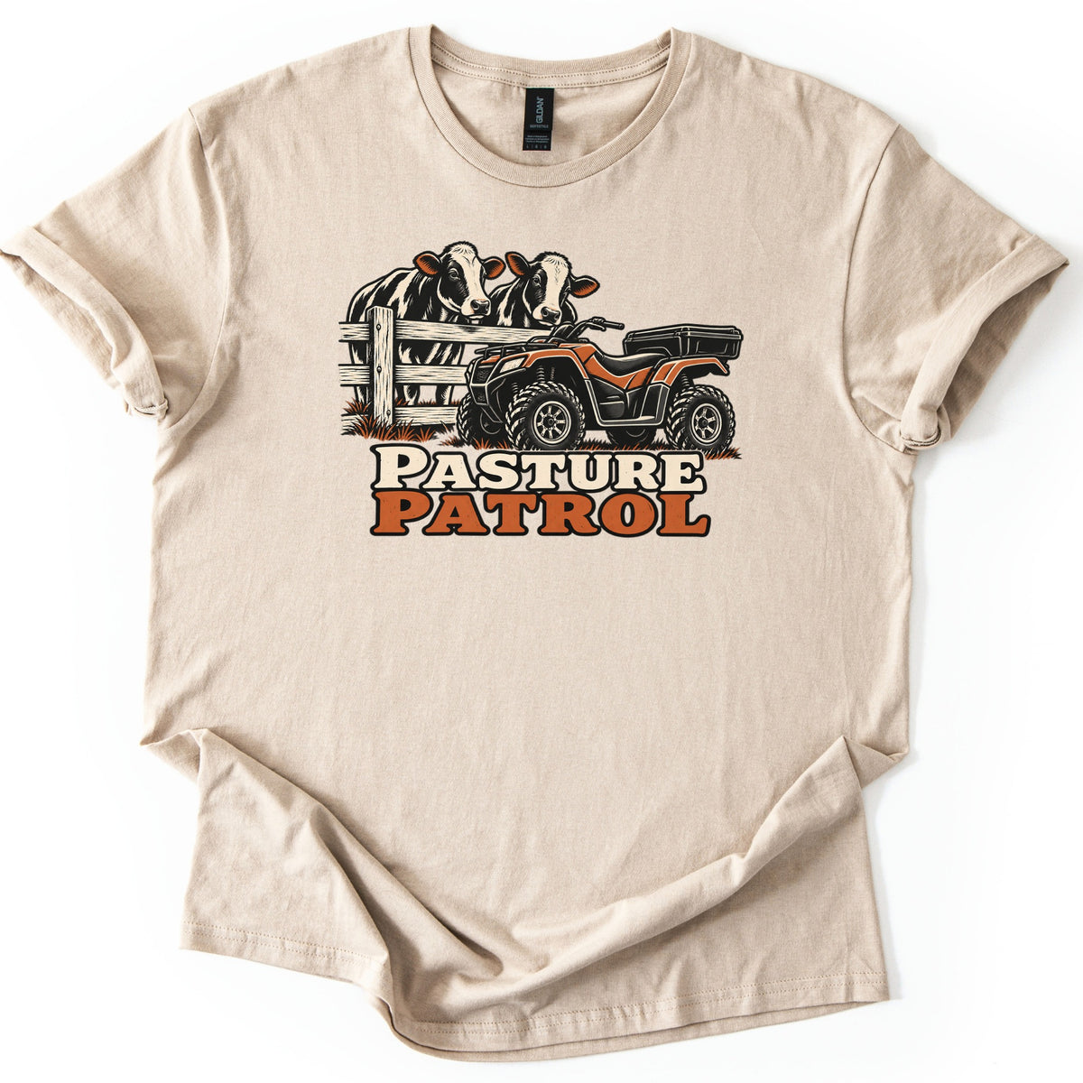 Pasture Patrol Comfort Colors T-shirt in Sand, laid flat. Two black and white cows look over a wooden fence beside an orange ATV with a rear cargo box, above the words Pasture Patrol. The shirt lies on a white background with its sleeves rolled.