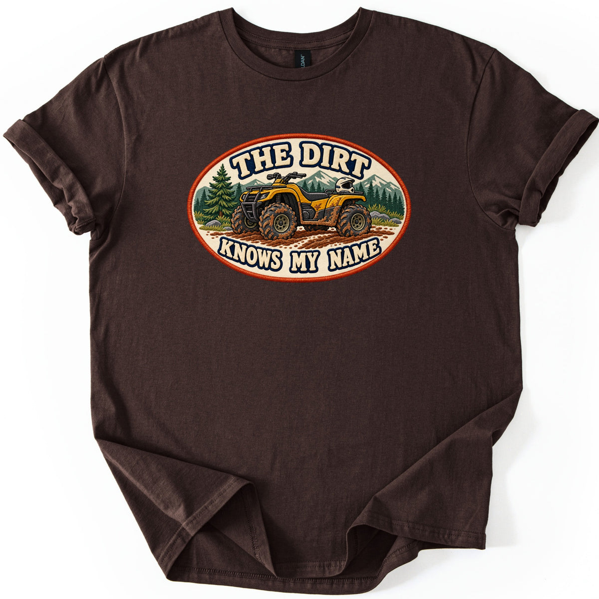 The Dirt Knows My Name Comfort Colors T-shirt in Dark Chocolate, laid flat. A cream oval patch shows a mud-splattered yellow ATV with a white helmet on its rack, pines and mountains behind, and the words THE DIRT KNOWS MY NAME. The shirt lies on a white surface with sleeves rolled.