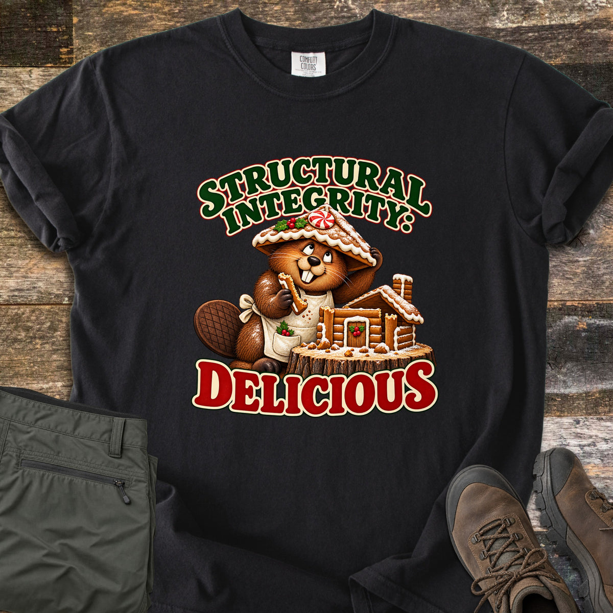 Structural Integrity: Delicious Comfort Colors T-shirt in Black, laid flat. A cartoon beaver wearing a gingerbread roof as a hat nibbles a cookie beside a gingerbread log cabin on a tree stump, between the words Structural Integrity: Delicious. Olive pants and brown boots lie on a wood floor.