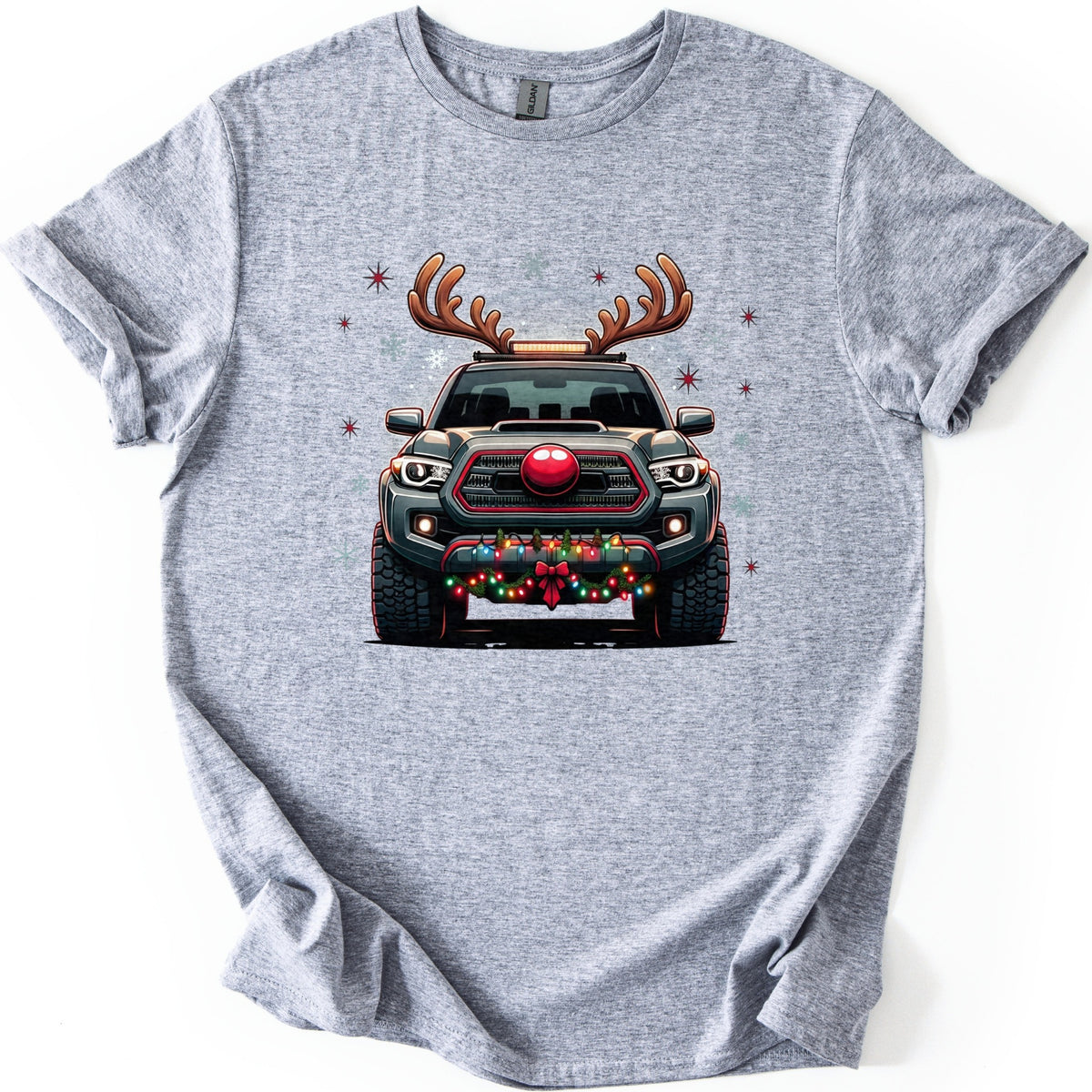 Santa Truck Comfort Colors T-shirt in Sport Grey, laid flat. A front-facing dark gray pickup truck with reindeer antlers, a round red nose on its grille, and colored lights with a red bow on the bumper, amid faint snowflakes and red stars. No words. White background.