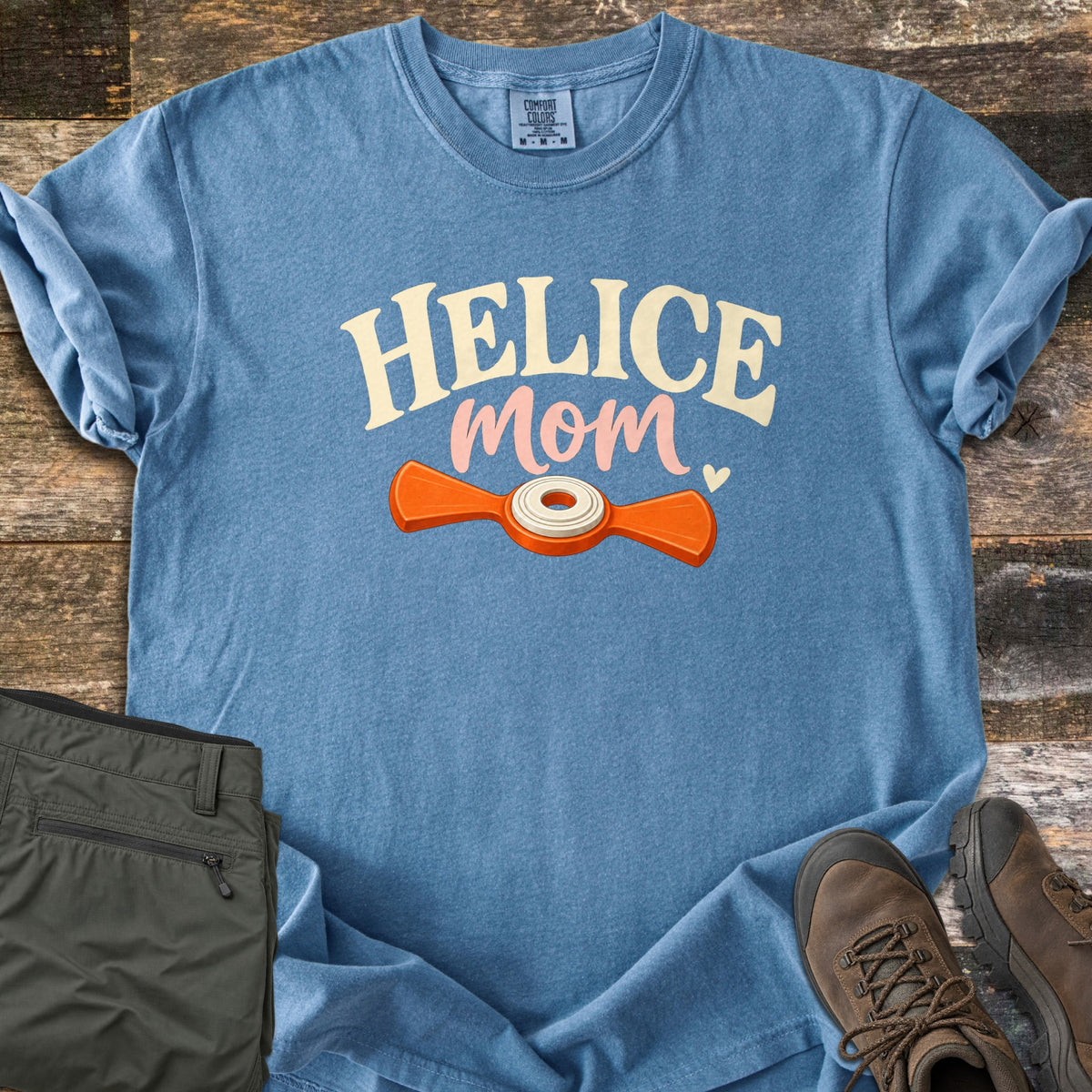 Helice Mom Comfort Colors T-shirt in Blue Jean, laid flat. HELICE in cream letters and mom in pink script with a small heart sit above an orange two-winged helice target with a white center. Folded olive pants and brown hiking boots rest beside it on weathered wood.