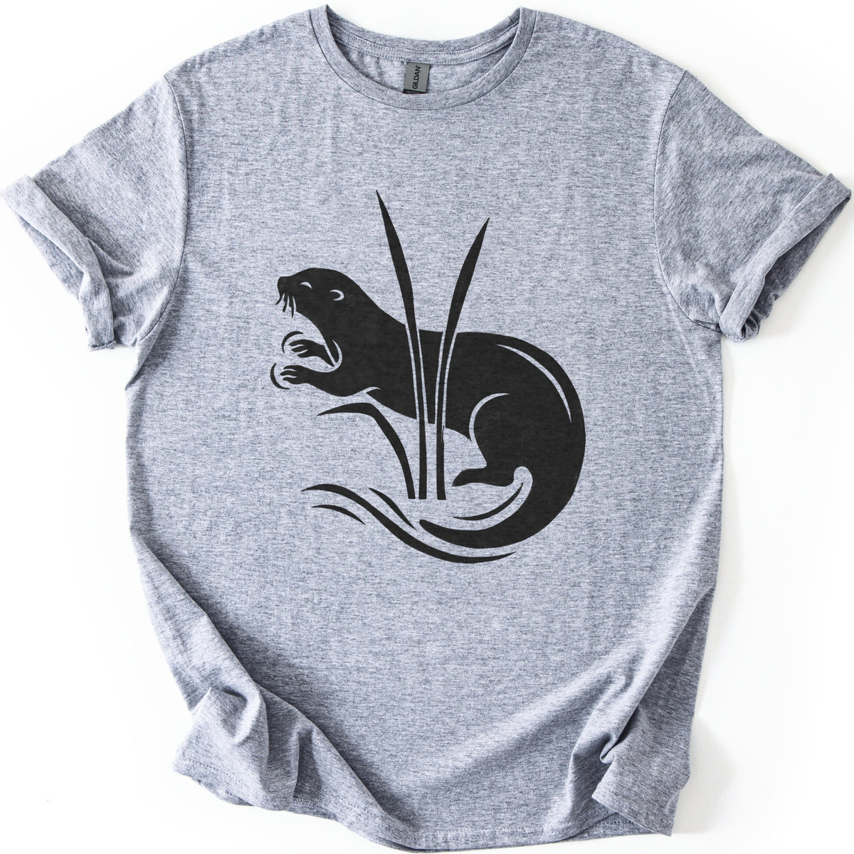 Otter In The Reeds Comfort Colors T-shirt in Sport Grey, laid flat. A black silhouette of an otter with open mouth, whiskers and raised paws curls behind two tall reeds above swirling water lines. No words. The shirt lies on a white surface with sleeves rolled.
