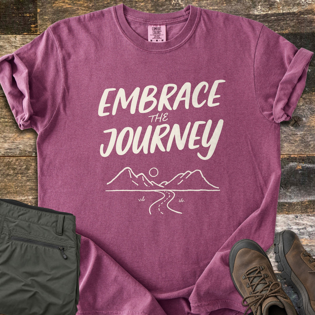 Embrace the Journey Comfort Colors T-shirt in Berry, laid flat. Cream brush lettering reads Embrace the Journey above a line drawing of mountains, a sun, and a winding road. Sleeves cuffed on weathered wood beside folded green pants and brown hiking boots.