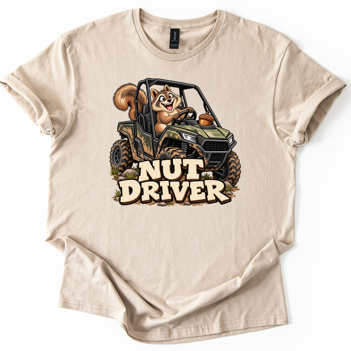 Nut Driver Comfort Colors T-shirt in Sand, laid flat. A grinning cartoon squirrel drives a muddy camo side-by-side UTV over rocks and grass, an acorn on the hood, above the words Nut Driver in cream letters. The shirt lies on a plain white background.