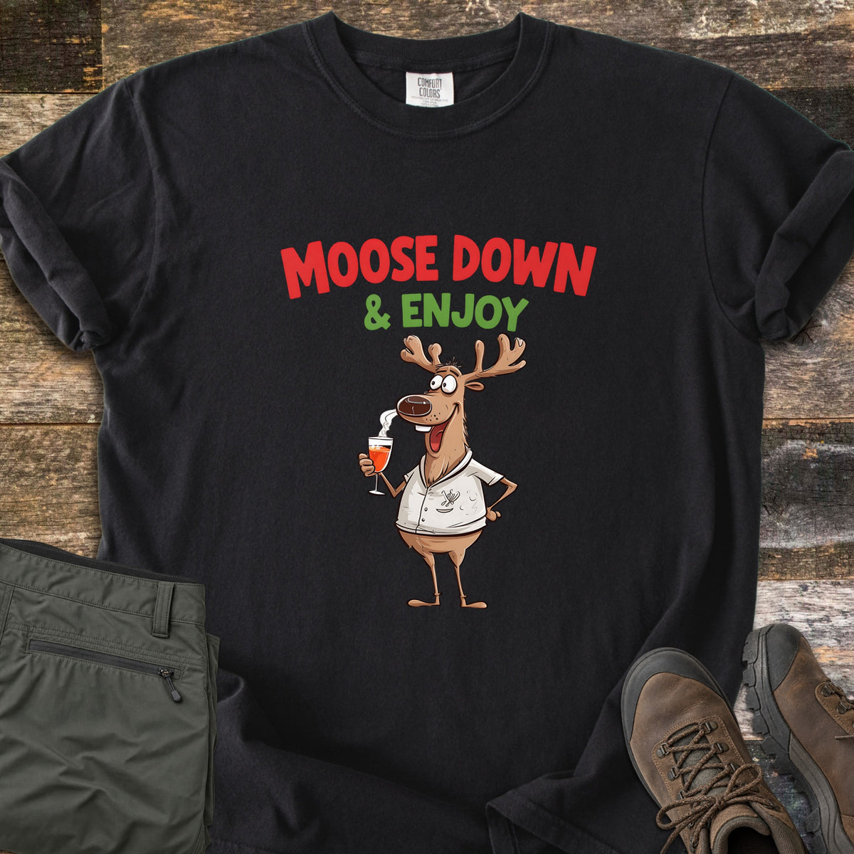Moose Down And Enjoy Comfort Colors T-shirt in Black, laid flat. A cartoon moose in a white shirt holds a glass of orange drink under the words Moose Down & Enjoy in red and green. Shown on rustic wood with folded olive pants and brown hiking boots.