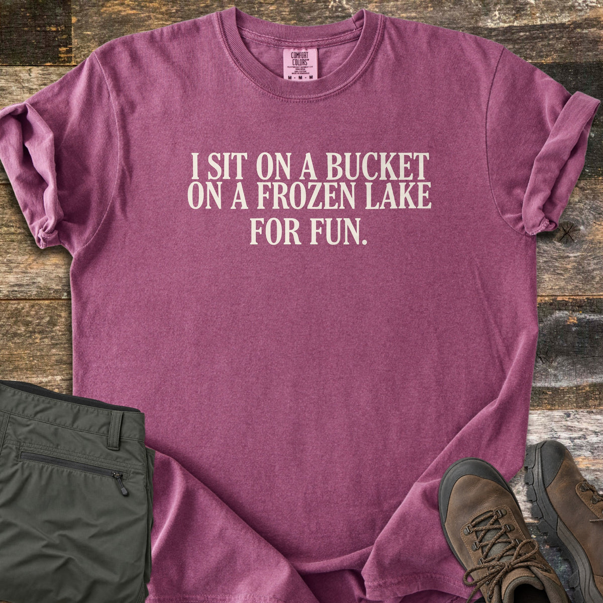 Bucket On A Frozen Lake Comfort Colors T-shirt in Berry, laid flat. Cream serif text reads I SIT ON A BUCKET ON A FROZEN LAKE FOR FUN. The shirt has rolled sleeves and lies on weathered wood planks beside folded olive hiking pants and brown hiking boots.