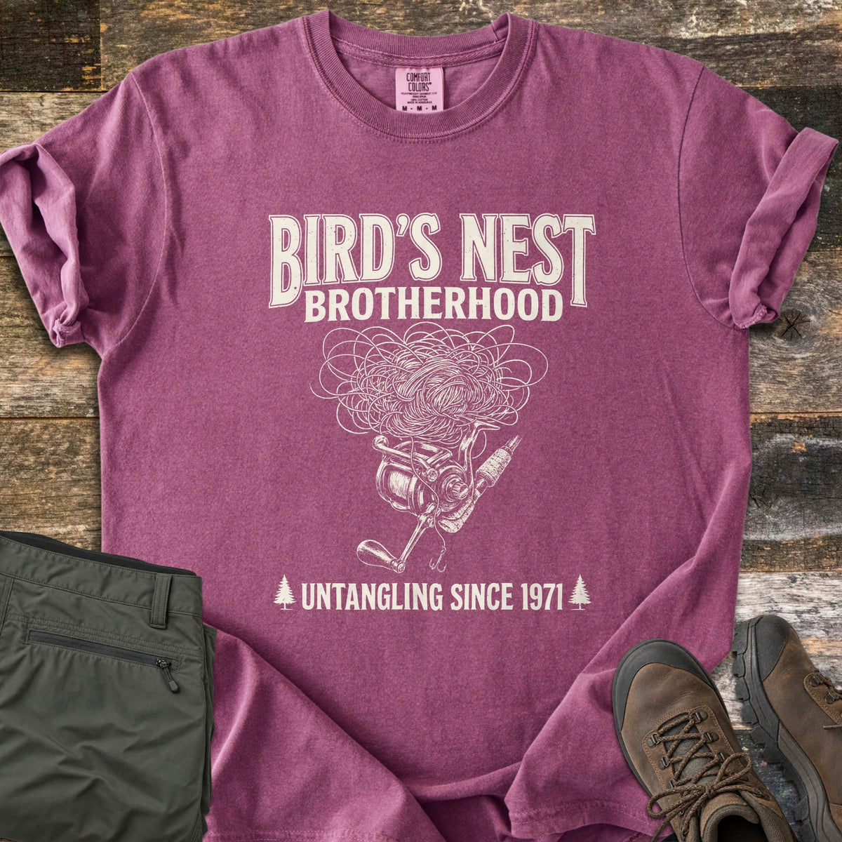 Birds Nest Brotherhood Comfort Colors T-shirt in Berry, laid flat. Cream sketch of a tangled line nest over a spinning reel, rod and hook, with Bird's Nest Brotherhood above and Untangling Since 1971 between pine trees below. Olive pants and hiking boots lie on old wood boards.