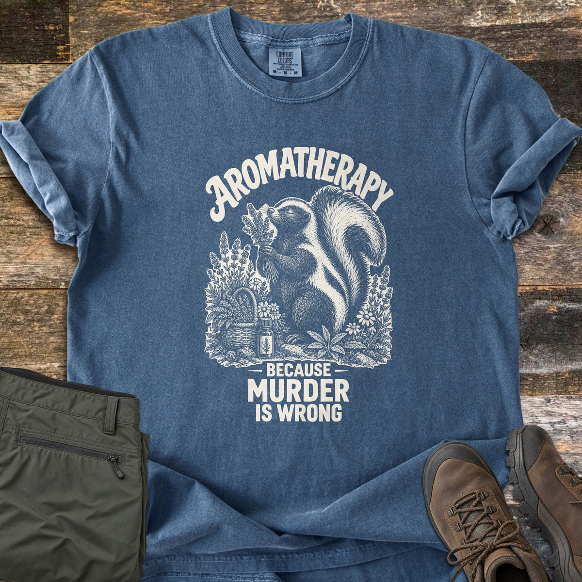 Aromatherapy Because Murder Is Wrong Comfort Colors T-shirt in Denim, laid flat. An engraving-style skunk sniffs a bundle of lavender among flowers, a basket and an oil bottle, in cream ink, with the words Aromatherapy, Because Murder Is Wrong. Olive pants and brown hiking boots lie on wood planks.