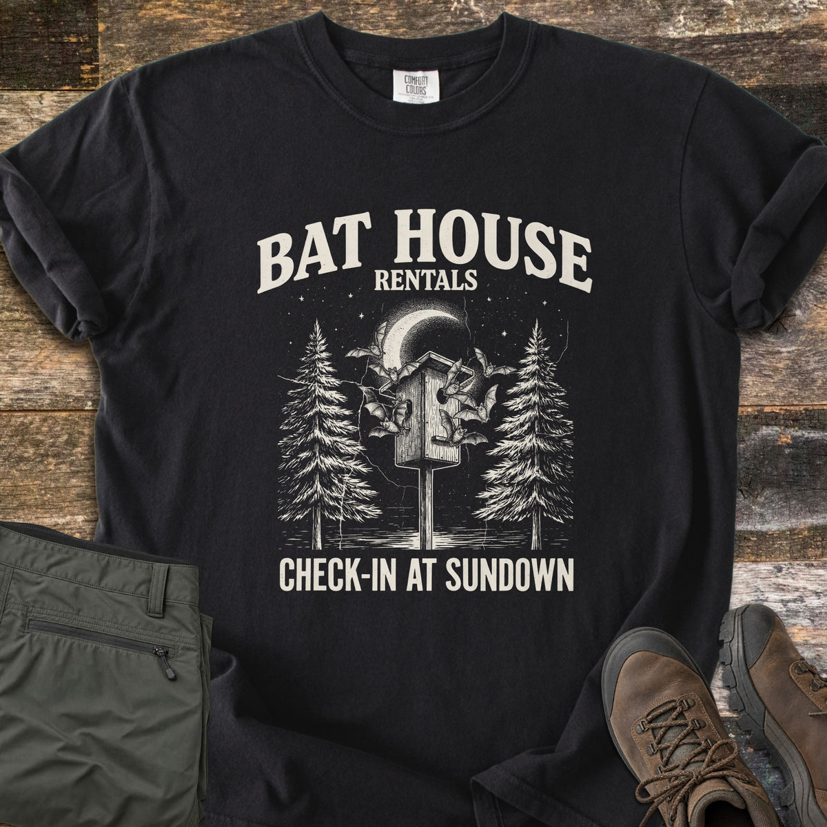 Bat House Rentals Comfort Colors T-shirt in Black, laid flat. Off-white print of a bat house on a post with bats flying around it under a crescent moon, between two pine trees, with the words Bat House Rentals and Check-In at Sundown. Olive pants and brown hiking boots lie on wood planks.