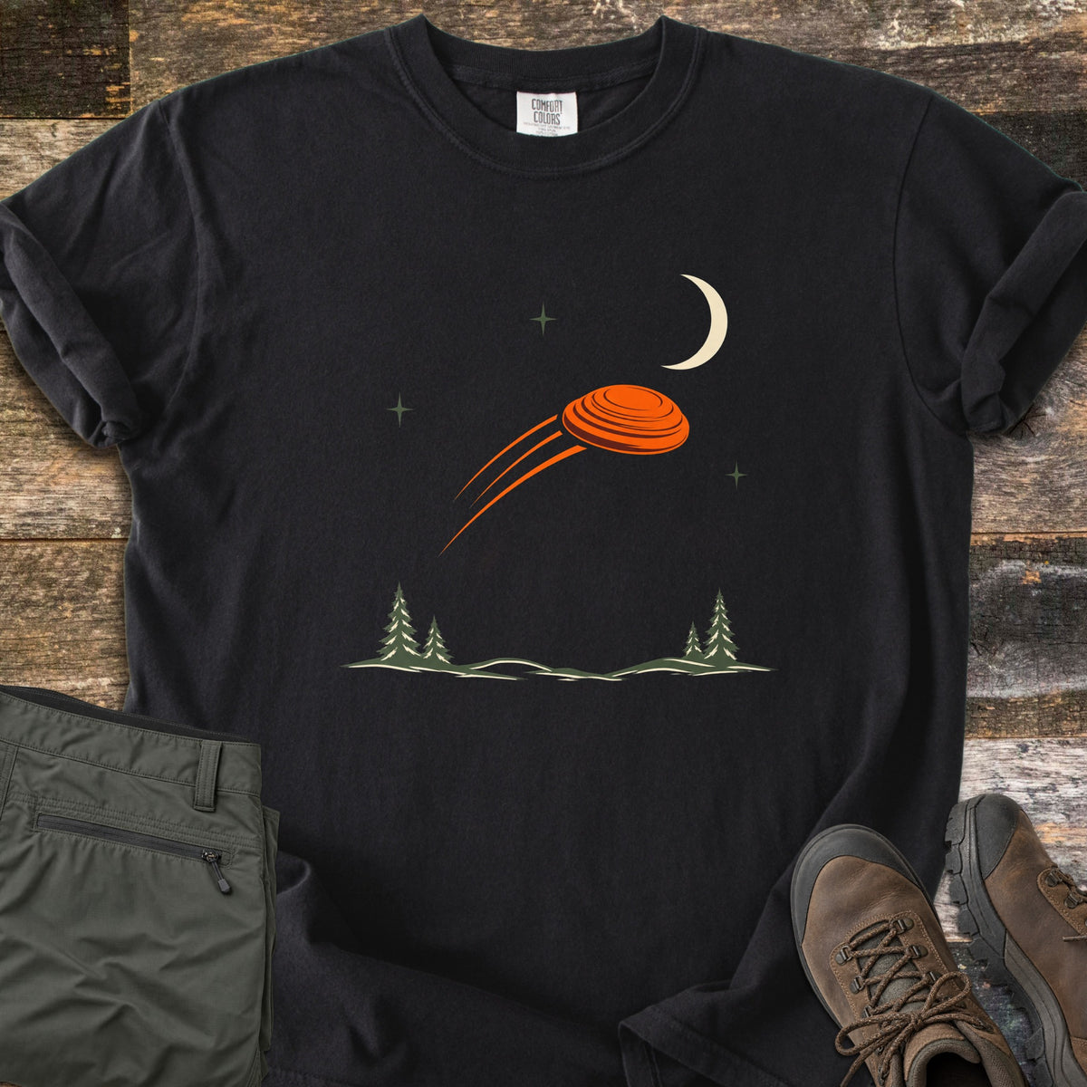 Moonrise Clay Comfort Colors T-shirt in Black, laid flat. An orange clay target streaks past a cream crescent moon and small stars over pine trees and snowy hills, with no words. Folded olive cargo pants and brown hiking boots lie beside it on weathered wood boards.