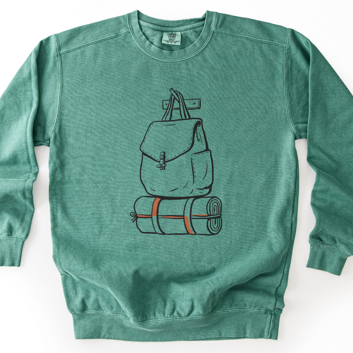 Backpack And Bedroll Comfort Colors crewneck sweatshirt in Light Green, laid flat. Black line art of a canvas backpack with a toggle clasp hanging by its straps from a wall peg above a rolled bedroll tied with orange straps. No words are printed. Shown on a white background.