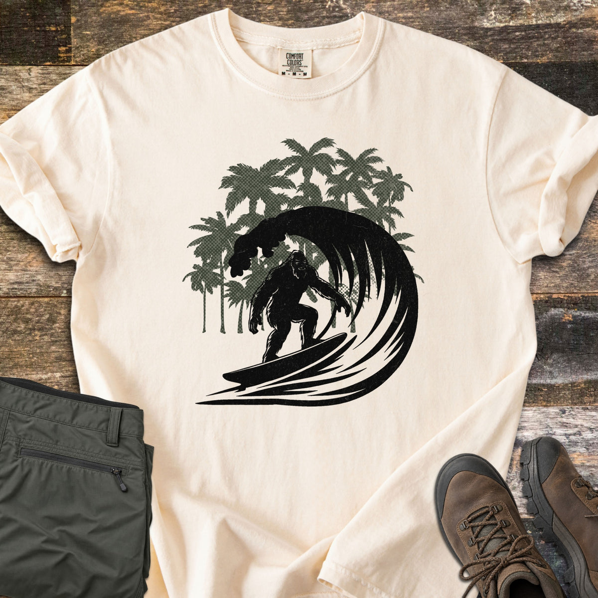 Surf Squatch Comfort Colors T-shirt in Ivory, laid flat. A black silhouette of Bigfoot surfing inside a curling wave, with green palm trees behind, and no words. The shirt lies on weathered wood with folded olive pants and brown hiking boots.