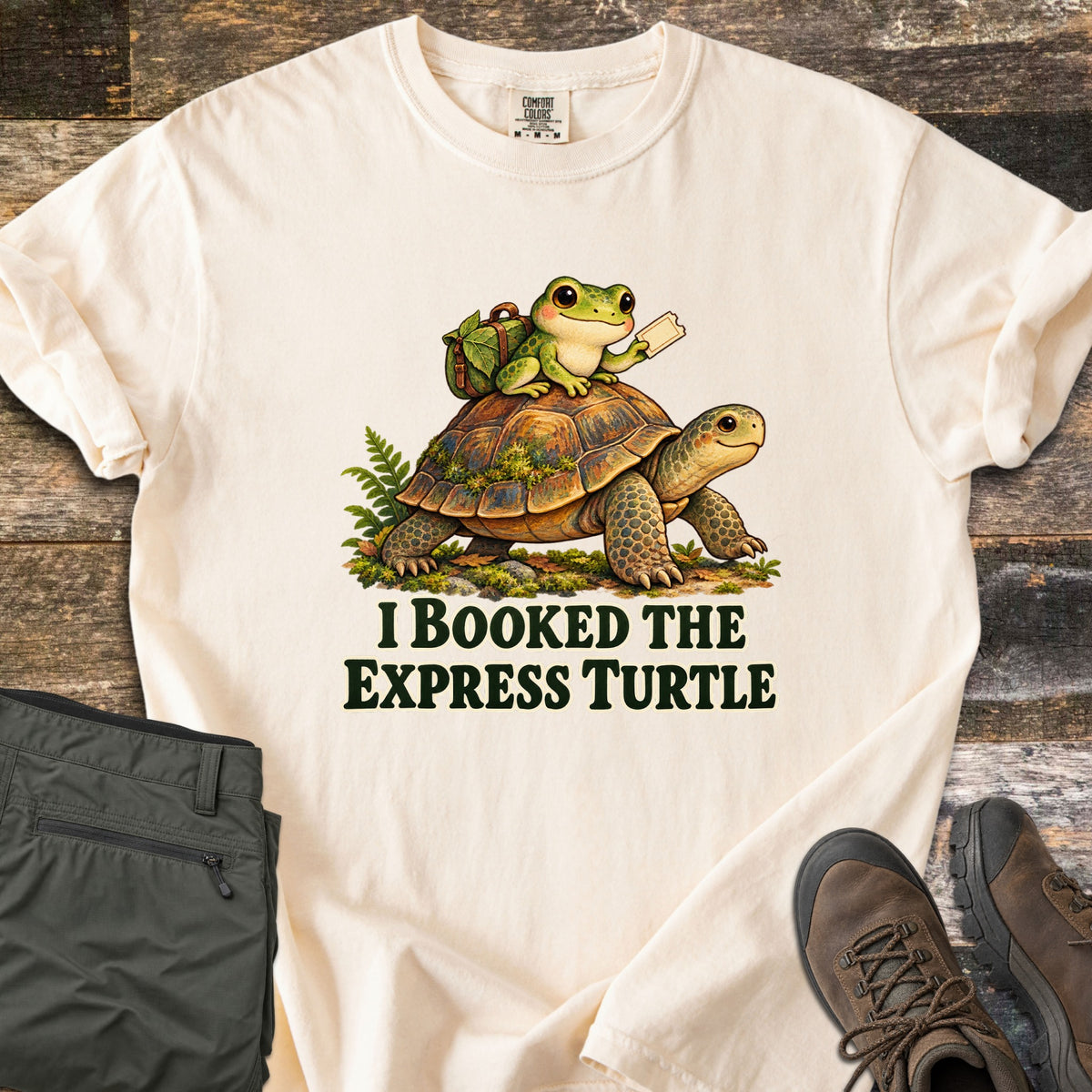 I Booked the Express Turtle Comfort Colors T-shirt in Ivory, laid flat. A cartoon frog with a leafy backpack holds a ticket while riding a mossy tortoise, above the words I Booked the Express Turtle in dark green. Shown on weathered wood with folded olive pants and brown hiking boots.