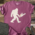 Lumber Squatch Comfort Colors T-shirt in Berry, laid flat. A distressed cream silhouette of Bigfoot walks while carrying a chainsaw with orange accents. The sleeves are rolled, with folded olive pants and brown hiking boots on rustic wood planks.