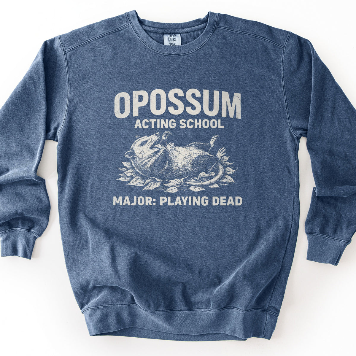 Opossum Acting School Comfort Colors crewneck sweatshirt in Denim, laid flat. Distressed off-white print of the words Opossum Acting School above an opossum lying on its back in leaves with its mouth open, and Major: Playing Dead below. Shown on a plain white background.