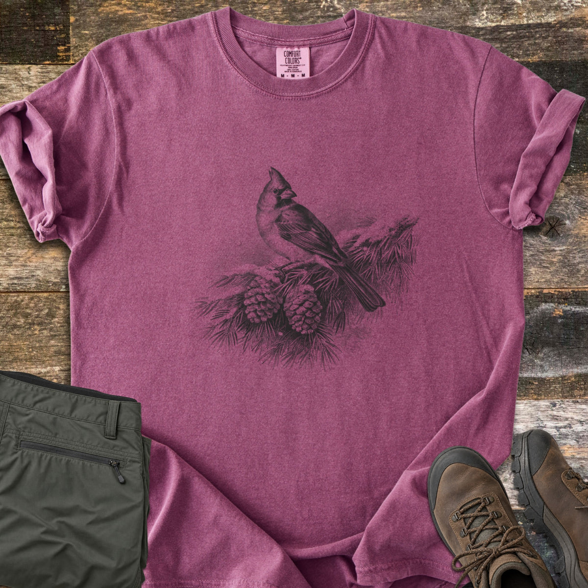 Cardinal On A Snowy Bough Comfort Colors T-shirt in Berry, laid flat. A grayscale drawing of a cardinal perched on a snow-covered pine branch with two pinecones, with no words. Sleeves rolled, on weathered wood planks beside olive hiking pants and brown hiking boots.