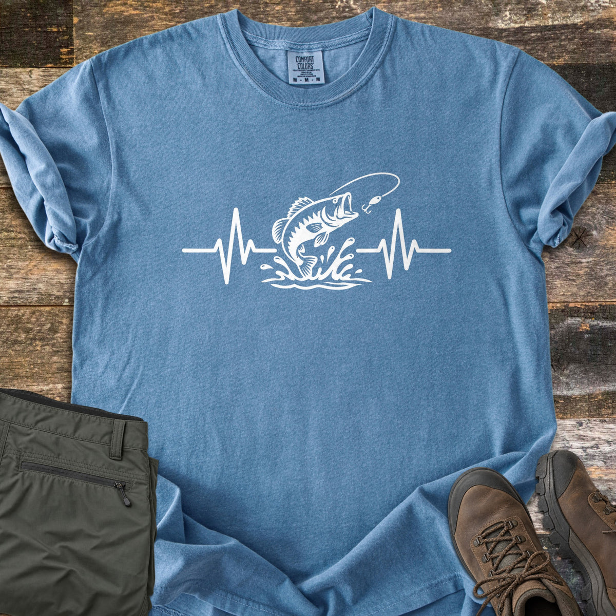 Fishing Heartbeat Comfort Colors T-shirt in Blue Jean, laid flat. A white print of a largemouth bass leaping from splashing water toward a fishing lure, set in the middle of a heartbeat line. Sleeves are rolled, on rustic wood with olive cargo shorts and brown hiking boots.