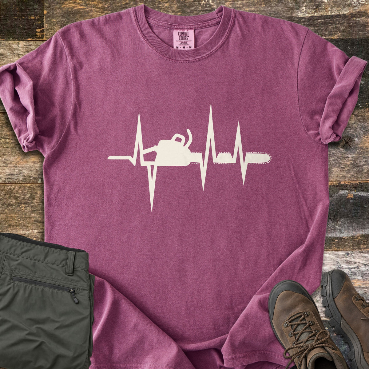 Chainsaw Heartbeat Comfort Colors T-shirt in Berry, laid flat. A cream heartbeat line runs across the chest with a chainsaw silhouette built into it, the bar and chain extending to the right. Folded olive cargo shorts and brown hiking boots sit on a weathered wood floor.