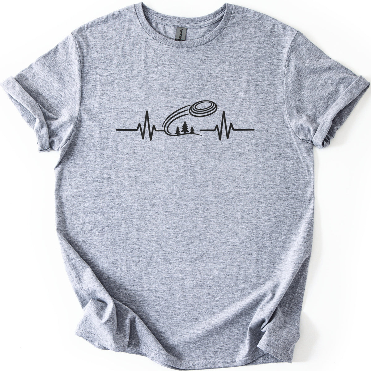 Trap Shooting Heartbeat Comfort Colors T-shirt in Sport Grey, laid flat. A black heartbeat line with a clay target flying above three pine trees on a hill, with no words. The shirt lies on a white background with its sleeves rolled.