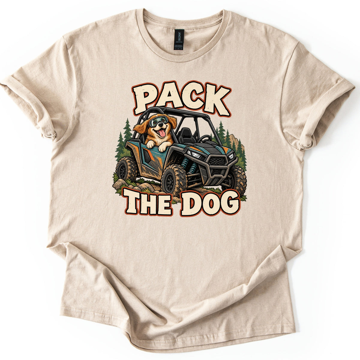 Pack the Dog Comfort Colors T-shirt in Sand, laid flat. A cartoon dog in goggles rides a muddy teal and black side-by-side UTV past pine trees, between the words PACK and THE DOG in cream letters. The shirt has cuffed sleeves on a white background.