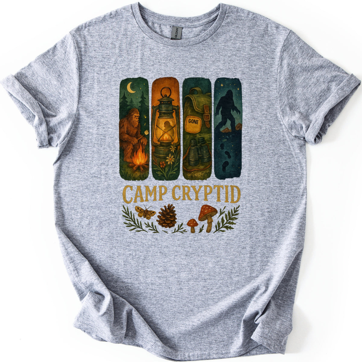 Camp Cryptid Big Foot Comfort Colors T-shirt in Sport Grey, laid flat. Four painted panels show Bigfoot at a campfire, a lit lantern, a backpack marked GONE, and Bigfoot's silhouette under stars, above the words Camp Cryptid and a moth, pinecone and mushrooms. Shown on a white background.