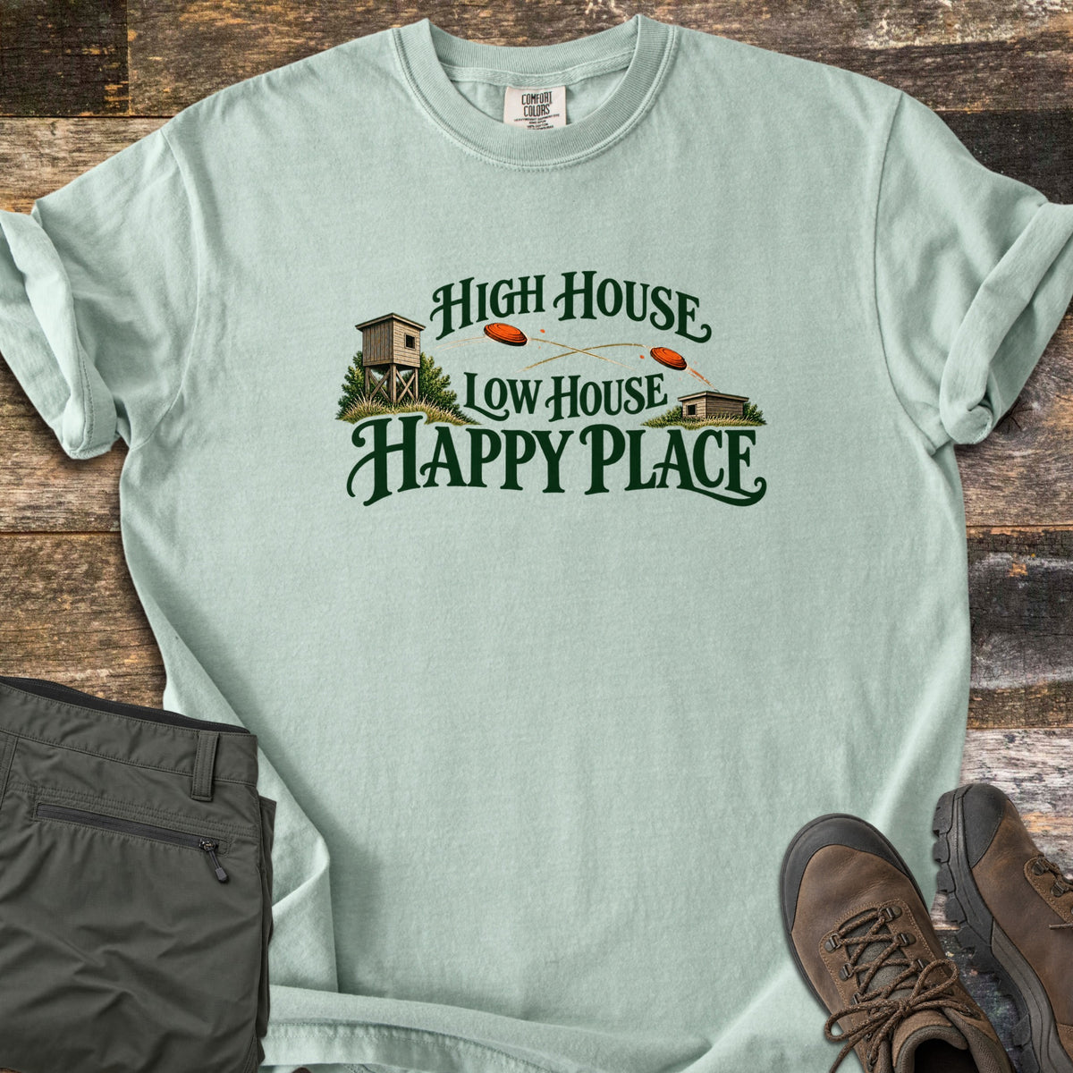High House Low House Happy Place Comfort Colors T-shirt in Bay, laid flat. Green lettering reads High House Low House Happy Place around two skeet houses with orange clay targets flying between them. Olive cargo pants and brown hiking boots lie beside it on weathered wood planks.