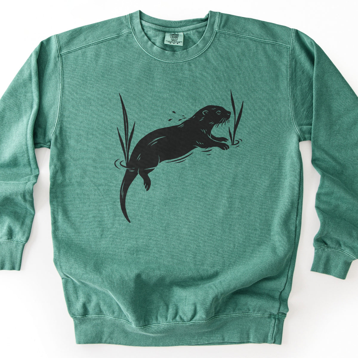 Swimming Otter Comfort Colors crewneck sweatshirt in Light Green, laid flat. A black block-print style otter swims with its head raised, splashing droplets, between two tall reeds with ripples around them. No words are printed. The sweatshirt lies on a white background.
