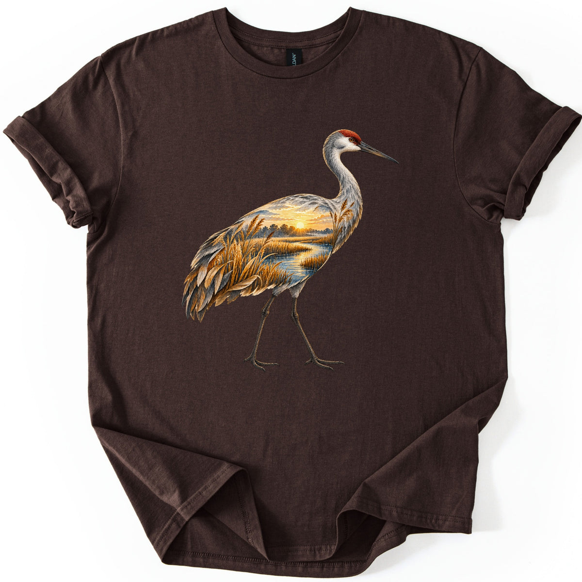 Crane Marsh Sunrise Comfort Colors T-shirt in Dark Chocolate, laid flat. A walking sandhill crane with a red crown whose body holds a marsh at sunrise, with golden reeds, a stream and the sun glowing over trees. No words. The shirt lies on a white background with cuffed sleeves.