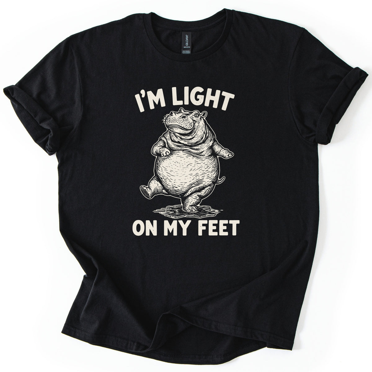 I'm Light On My Feet Comfort Colors T-shirt in Black, laid flat. A cream etched-style drawing of a round hippo standing upright on one foot, mid-step, between the words I'M LIGHT above and ON MY FEET below. The shirt has rolled sleeves on a white background.