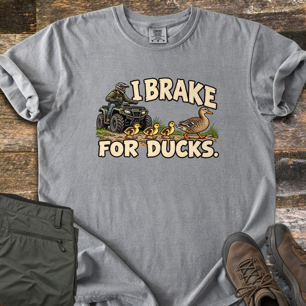 I Brake for Ducks Comfort Colors T-shirt in Grey, laid flat. A rider in a helmet on a green ATV waits as a mother duck leads her ducklings across a dirt path, under the words I Brake For Ducks. Olive pants and brown hiking boots lie beside it on weathered wood planks.