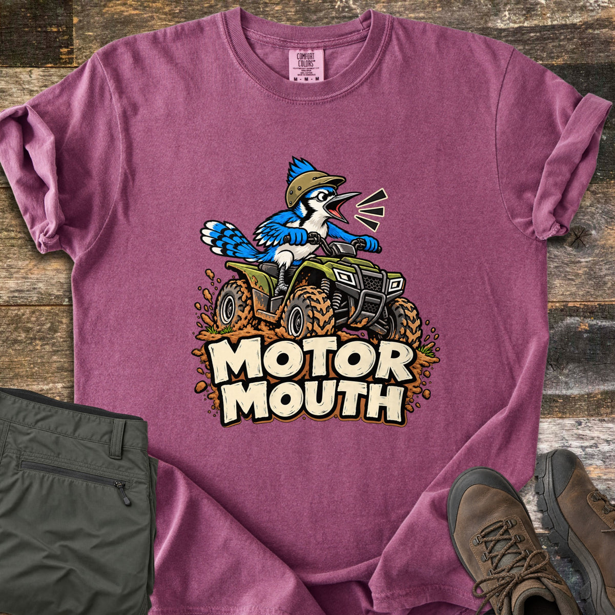 Motor Mouth Comfort Colors T-shirt in Berry, laid flat. A shouting cartoon blue jay in a tan helmet drives a muddy green four-wheeler above the words Motor Mouth. The sleeves are rolled, with folded olive pants and brown hiking boots on weathered wood.