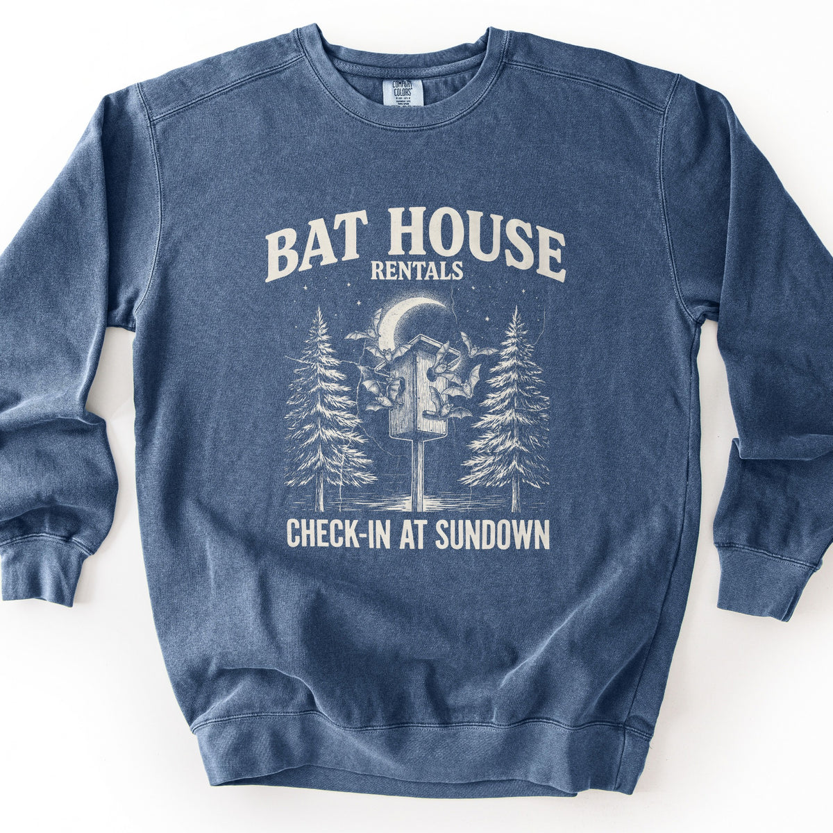 Bat House Rentals Comfort Colors crewneck sweatshirt in Denim, laid flat. Cream print of a wooden bat house on a post with bats flying around it, a crescent moon, stars and two pine trees, under the words Bat House Rentals and above Check-In at Sundown. Shown on a white background.