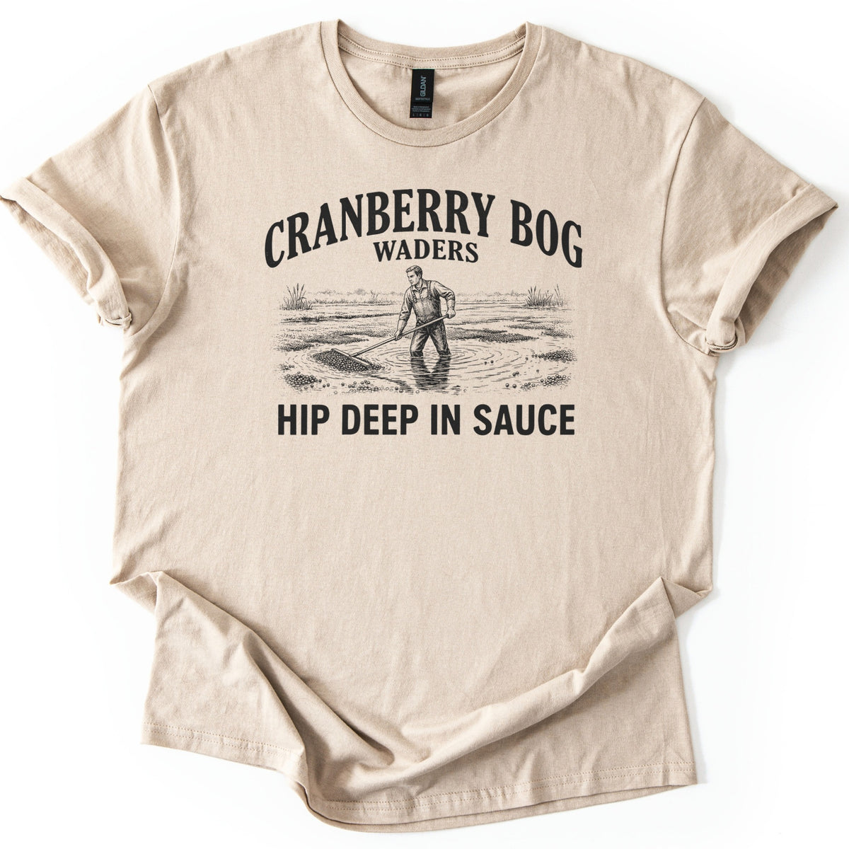 Cranberry Bog Waders Comfort Colors T-shirt in Sand, laid flat. A black etched drawing of a man in overalls standing hip deep in a flooded bog, raking floating cranberries, under the words Cranberry Bog Waders and above Hip Deep in Sauce. Sleeves cuffed, on white.