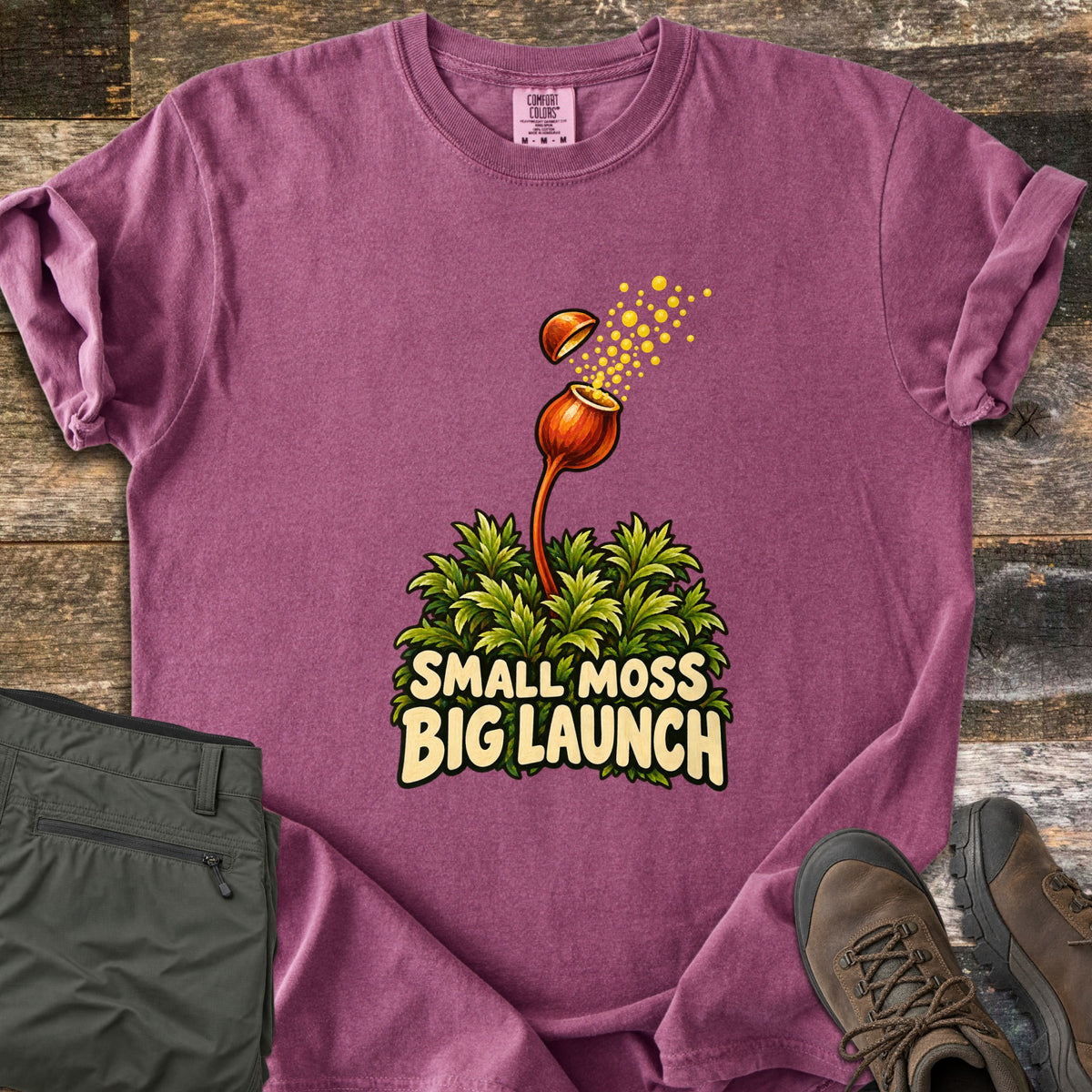 Small Moss Big Launch Comfort Colors T-shirt in Berry, laid flat. Yellow spores burst from an orange moss capsule whose lid pops off, rising on a stalk from green moss over the words Small Moss Big Launch. Olive hiking pants and brown boots lie beside it on wood planks.