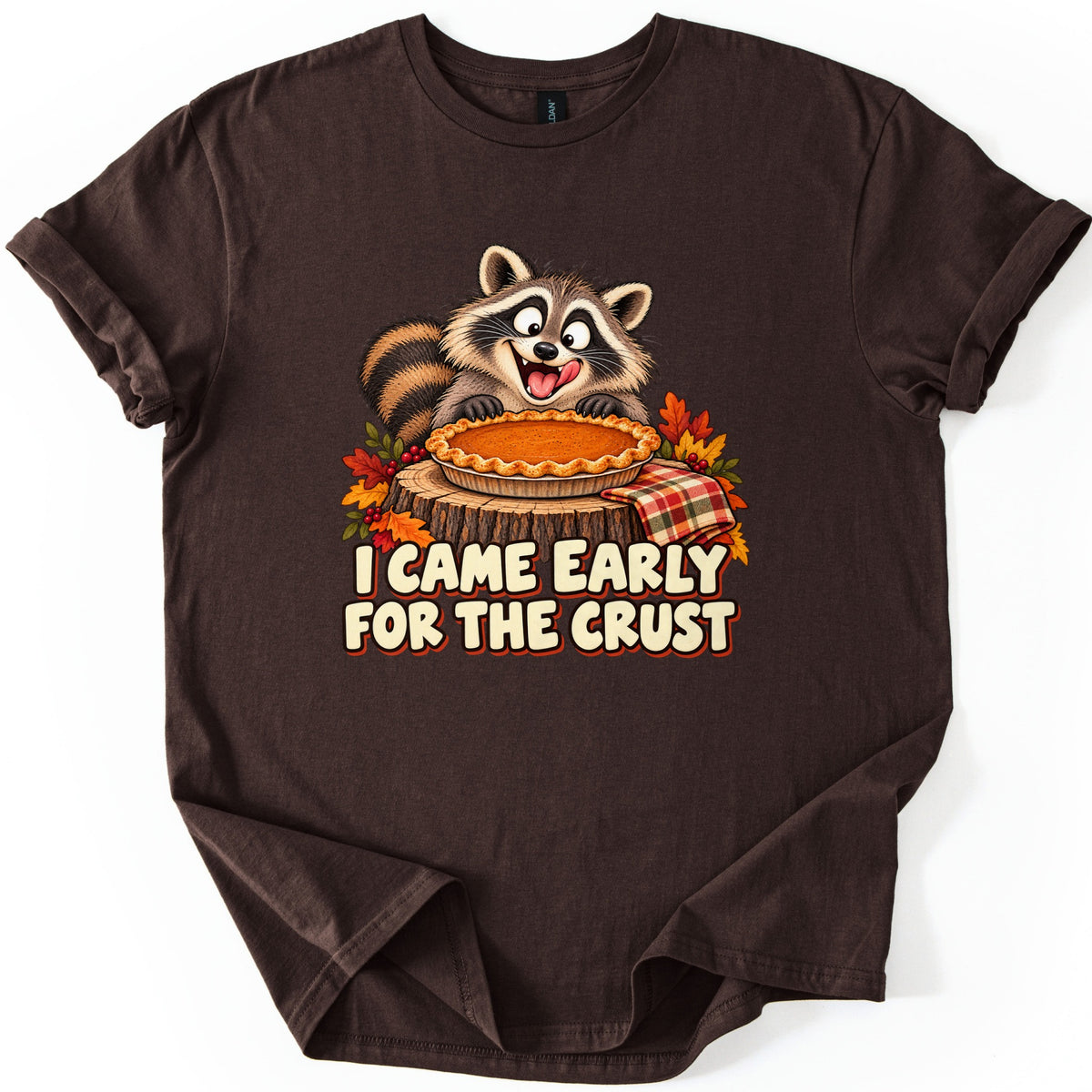 I Came Early for the Crust Comfort Colors T-shirt in Dark Chocolate, laid flat. A wide-eyed cartoon raccoon leans over a pumpkin pie set on a tree stump with autumn leaves and a plaid cloth, above the words I CAME EARLY FOR THE CRUST. Sleeves are cuffed on a white background.