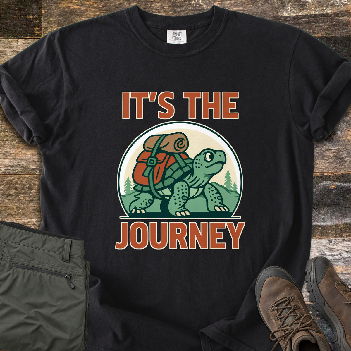 Slow Turtle Hiker Journey Comfort Colors T-shirt in Black, laid flat. A cartoon turtle carrying a red backpack and bedroll walks past pine trees and hills between the words It's the Journey. Olive hiking pants and brown hiking boots sit beside it on rustic wood planks.