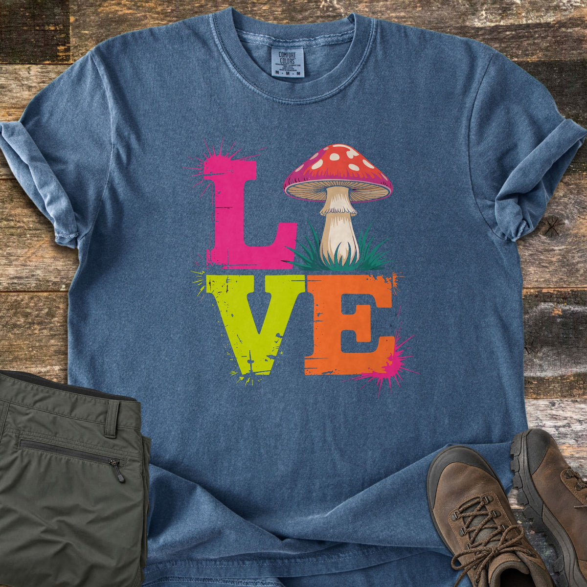 Mushroom Love Comfort Colors T-shirt in Denim, laid flat. The word LOVE in distressed pink, lime green and orange letters with paint splatters, and a red spotted mushroom in grass as the O. Laid on weathered wood with olive pants and brown hiking boots.