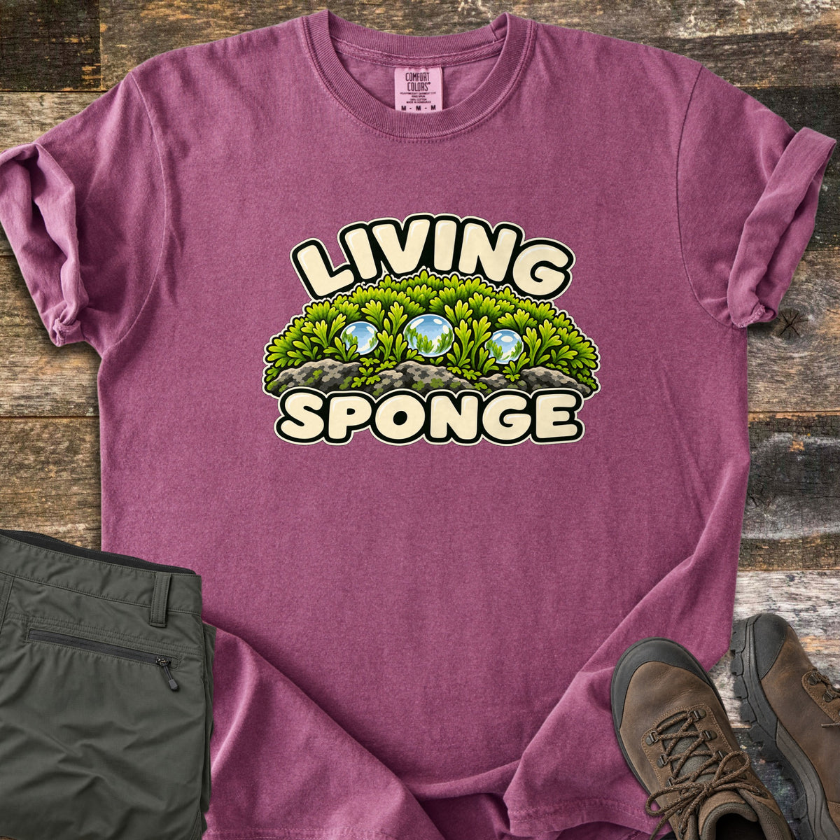 Living Sponge Comfort Colors T-shirt in Berry, laid flat. A cartoon mound of bright green moss on gray rock, holding three clear water droplets, between the words Living Sponge. Olive hiking pants and brown leather hiking boots lie beside it on weathered wood.