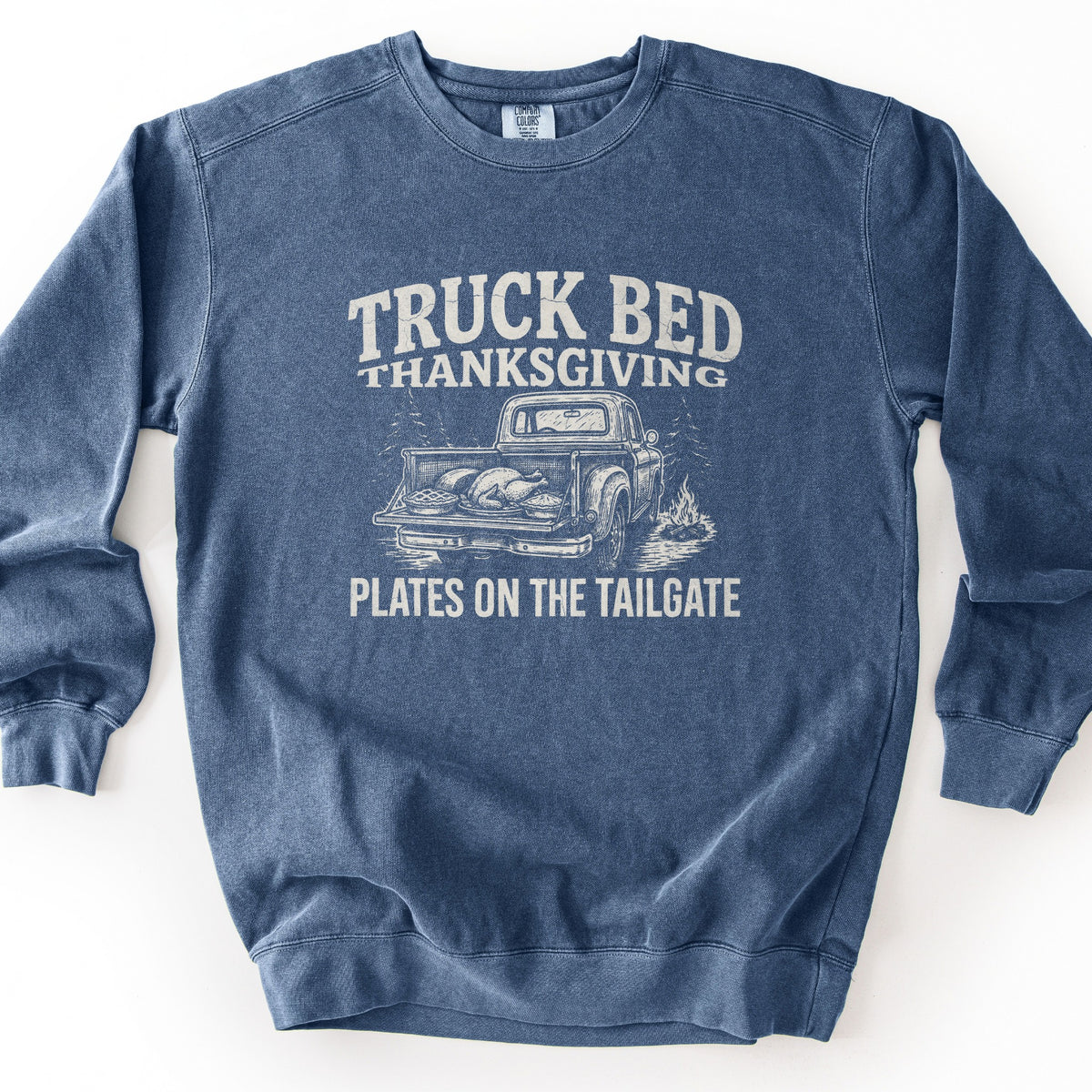 Truck Bed Thanksgiving Comfort Colors crewneck sweatshirt in Denim, laid flat. Distressed off-white print reads Truck Bed Thanksgiving above a sketched vintage pickup with a roast turkey and pies in its open bed, pine trees and a campfire, and the words Plates on the Tailgate. Shown on a white background.