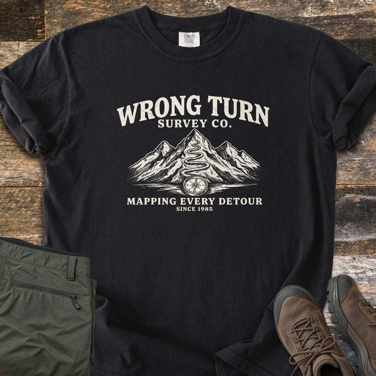 Wrong Turn Survey Co Comfort Colors T-shirt in Black, laid flat. Cream print of the words Wrong Turn Survey Co. above snowy mountains with a winding trail leading to a compass, then Mapping Every Detour, Since 1985. Shown on wood boards with olive pants and brown hiking boots.