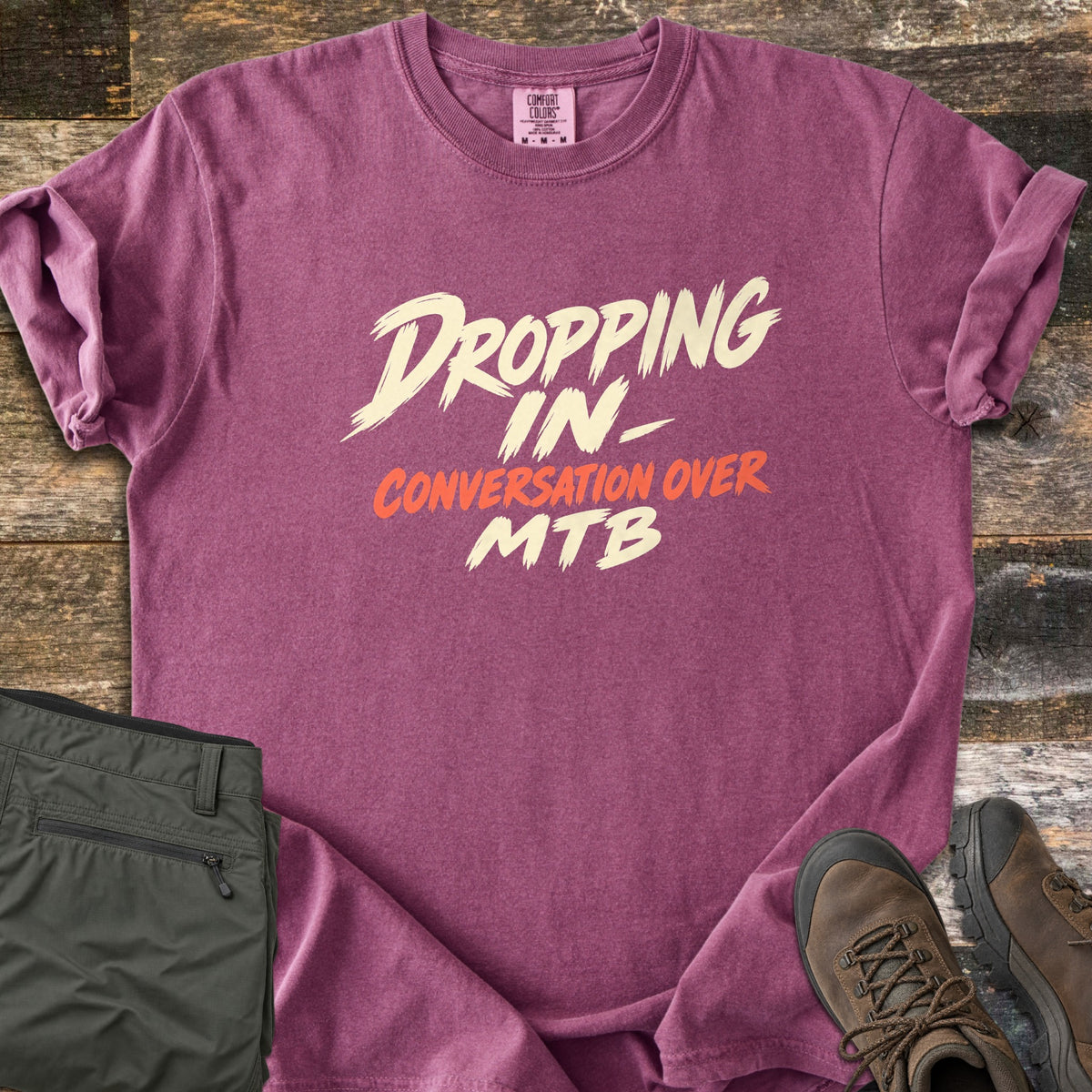Dropping In Conversation Over Comfort Colors T-shirt in Berry, laid flat. Cream brush-stroke letters read Dropping In- and MTB, with Conversation Over in orange-red between them. Rolled sleeves rest on weathered wood planks near folded olive pants and brown hiking boots.